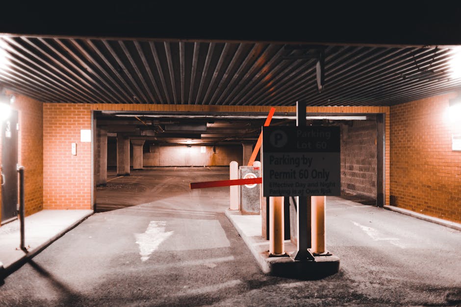 Photo of an underground parking garage entrance with red brick walls and a black corrugated ceiling. A sign mounted on a metal post indicates parking lot 60 and parking by permit only, with text partially visible. Two orange and black traffic barriers are positioned in front of the entrance, with one arm extending across the opening, likely to control access. In front of the barriers, a pavement area shows a white arrow painted on the ground pointing inward, guiding vehicles or pedestrians. The interior of the parking garage is dimly lit, with concrete pillars and a concrete ceiling visible in the background, creating a spacious, utilitarian environment suitable for vehicle parking and access control during a home relocation or moving process. This scene is associated with Man with Van Thamesmead, providing moving and removal services, highlighting the logistical environment for furniture transport and packing in an urban setting.