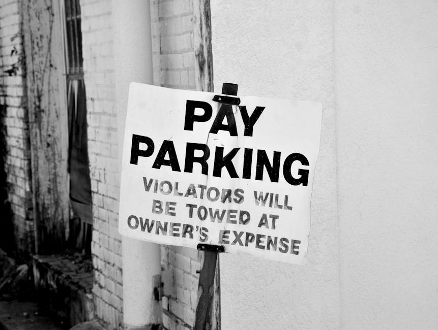 A black-and-white photograph depicting a rectangular 'Pay Parking' sign attached to a weathered wooden post, positioned next to a brick wall and a textured concrete surface. The sign features bold black text on a white background, with the message 'Pay Parking' prominently at the top, followed by smaller text stating 'Violators will be towed at owner's expense.' The sign is slightly tilted and secured with metal clips. The setting appears to be outside a building, possibly in an urban area, with visible signs of aging on the wall and post. This image relates to moving logistics and parking regulations relevant to house removals, similar to services offered by Man with Van Thamesmead, especially concerning parking permits and avoiding fines during home relocation or furniture transport.