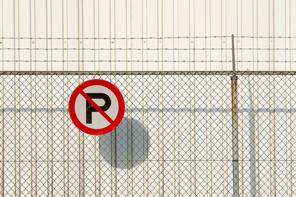 Photo of an underground parking garage entrance with red brick walls and a black corrugated ceiling. A sign mounted on a metal post indicates parking lot 60 and parking by permit only, with text partially visible. Two orange and black traffic barriers are positioned in front of the entrance, with one arm extending across the opening, likely to control access. In front of the barriers, a pavement area shows a white arrow painted on the ground pointing inward, guiding vehicles or pedestrians. The interior of the parking garage is dimly lit, with concrete pillars and a concrete ceiling visible in the background, creating a spacious, utilitarian environment suitable for vehicle parking and access control during a home relocation or moving process. This scene is associated with Man with Van Thamesmead, providing moving and removal services, highlighting the logistical environment for furniture transport and packing in an urban setting.