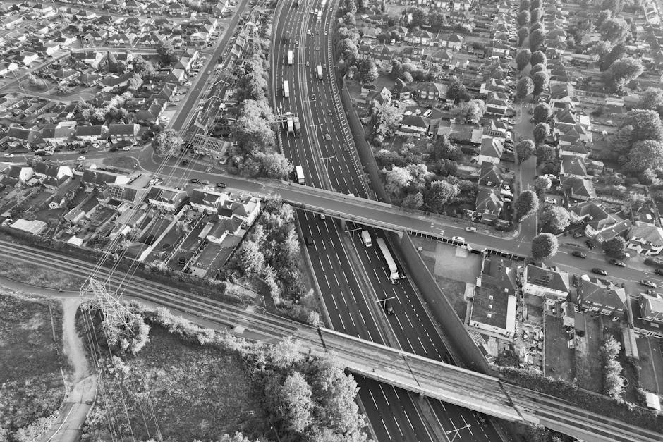 An aerial black-and-white photograph showing a busy suburban area with residential houses and streets in Thamesmead, SE28. The image features a major multi-lane road running vertically through the scene, with several vehicles including trucks and cars traveling along it. Over this main road, a smaller bridge or overpass crosses, supported by concrete pillars, facilitating the movement of traffic above the lower road. Adjacent to the road are railway tracks with electrical wiring and pylons running parallel, indicating the proximity of rail transport to the residential zones. Surrounding the roads are numerous houses, many with pitched roofs, gardens, and trees, typical of a suburban neighbourhood. The area appears well-developed with a mixture of commercial and residential buildings, and the lighting suggests daytime conditions. This setting exemplifies urban and suburban infrastructure, relevant to house removals and moving logistics, where careful planning of transport routes is essential, which [COMPANY_NAME], Man with Van Thamesmead, can assist with during home relocation or furniture transport services.