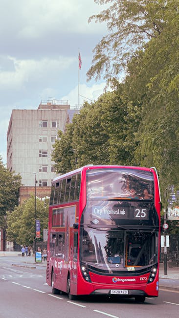 A double-decker bus, painted in bright pink, is driving along a tree-lined street in Thamesmead. The bus displays the route number 25 and the destination 'City Thameslink' on the front. Behind the bus, there are tall buildings with a partly cloudy sky above. The street is lined with green trees whose foliage extends over the pavement, providing shade. The bus is part of a moving process, possibly indicating transport logistics for home relocation or furniture transport. The scene captures the urban environment associated with house removals, with the bus as a key element in relocation services offered by Man with Van Thamesmead, situated in a context that supports effective packing and moving operations across Thamesmead's SE28 area.