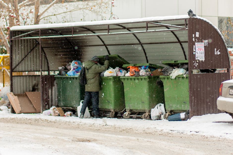 An outdoor scene in an urban area showing multiple waste and recycling bins, including a large grey bin filled with mixed paper and cardboard packaging, some of which are overflowing and leaning open. Adjacent to the grey bin are black recycling bags, a red bin, and additional cardboard boxes and plastic waste on the ground nearby. The area is paved with bricks, with a metal railing separating the waste collection point from the street, where a silver car is parked. In the background, a building with a blue metal staircase and scaffolding can be seen, along with storefronts featuring logos and signs indicating recycling zones. The image is taken during daylight, under natural lighting, capturing the typical scene of home relocation or moving logistics involving packing and disposal of household materials, which Man with Van Thamesmead offers as part of their removals service.