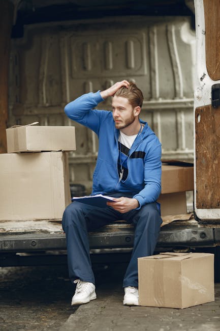 A man wearing a blue long-sleeve shirt, black quilted vest, black pants, white sneakers, and a grey cap stands next to the open rear door of a white moving van parked on a brick driveway outside a res