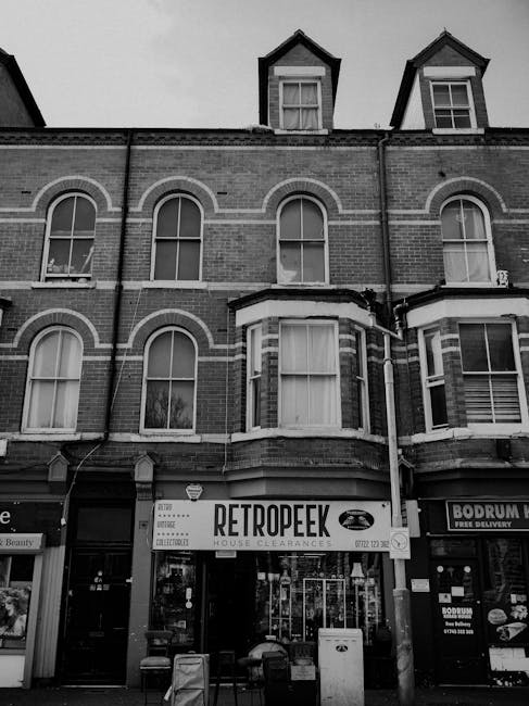 Black and white photograph of a multi-storey residential building with three visible floors, situated on a street with commercial units at ground level. The building features brickwork with arched and rectangular windows, some with curtains or blinds visible. On the ground floor, there are two store shopfronts; the left one has signage reading 'RETROPEEK HOUSE CLEARANCES' indicating a house clearance service, while the right one advertises food delivery. In front of the shops, on the pavement, there are various items associated with house removals such as a large cardboard box, packing materials, and a trolley, suggesting recent or ongoing furniture transport or packing activities. Part of a van or delivery vehicle is slightly visible on the right side, parked close to the building, with the scene capturing a typical urban setting for home relocation or furniture moving services. The environment is illuminated by natural light, and the overall scene emphasizes the process of packing, loading, or house clearance in an urban street context.