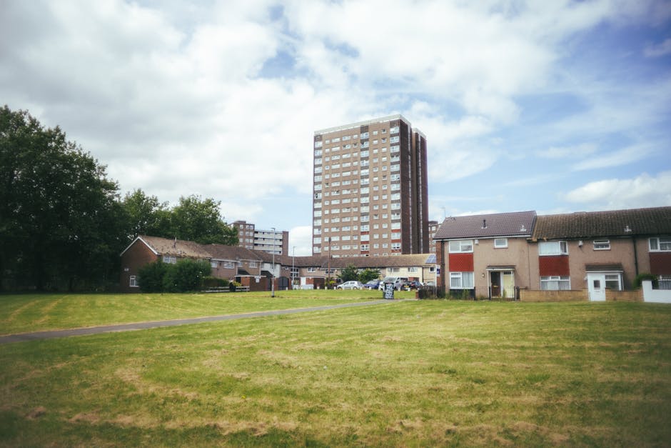 A wide view of a residential exterior scene with a large, grassy open area in the foreground, featuring a narrow paved footpath running across the grass. Behind the grass, there are several small, single-story houses with brown and beige exteriors, some with white doors and window frames, and pitched roofs covered in dark shingles. To the right, a white van with its rear doors open is visible, with partially loaded packing boxes and furniture covered in protective wrapping, likely part of a home relocation or furniture transport process. Further in the background, taller apartment buildings with multiple floors, beige and brown façades, and numerous windows loom over the neighborhood, creating a contrast between the low-rise homes and high-rise apartments. The sky above is partly cloudy with patches of blue, indicating daytime, and the scene suggests a relocation or moving logistics environment, with Man with Van Thamesmead providing professional removals services.
