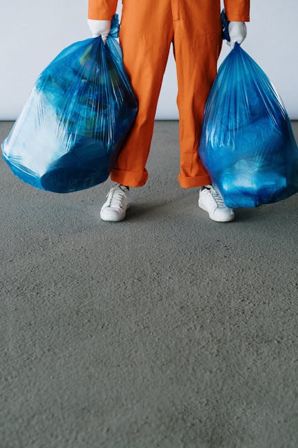 A person wearing orange work pants and white sneakers stands on a gray indoor floor, holding two large blue plastic garbage bags filled with waste or bulky items, suggesting a home relocation or removal process. The individual’s hands, covered with gray gloves, grip the bags at each side, with the bags appearing slightly stretched and full. The background shows a plain, light-colored wall with natural lighting, and part of the person's upper body is obscured, focusing on the action of handling waste materials. This scene reflects a typical furniture or waste removal activity often associated with property clearing or moving logistics, emphasizing the importance of proper handling and transport of bulky waste during house removals, as provided by Man with Van Thamesmead.