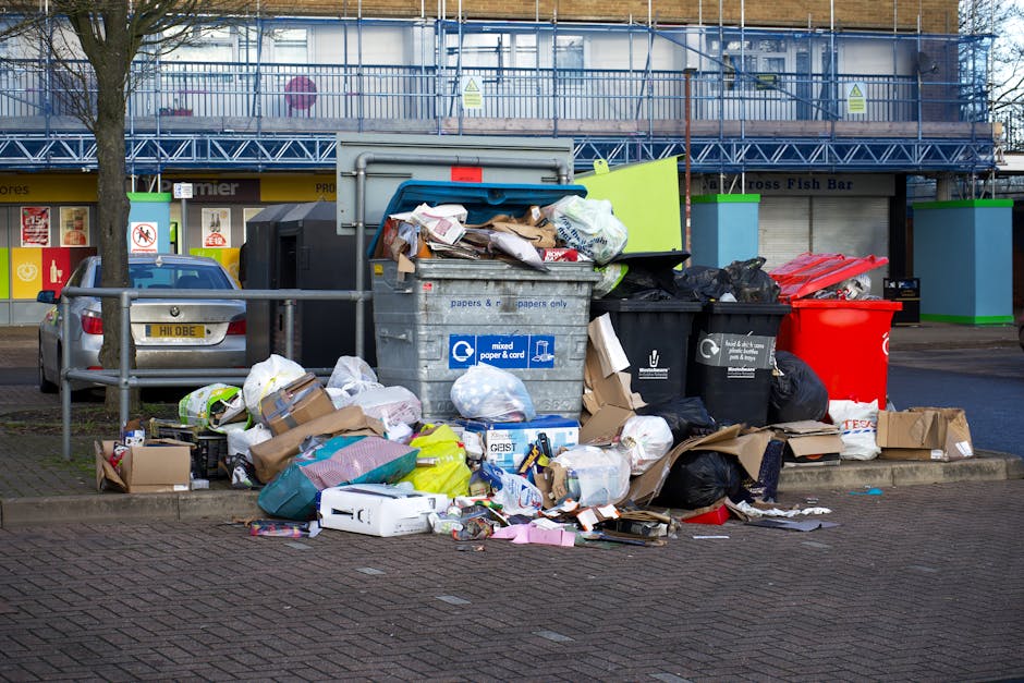 A large outdoor rubbish bin filled with mixed paper, cardboard, plastic bags, and various packaging materials, with some items spilling onto the surrounding brick pavement. Several black and red waste bags are placed next to the bin, along with flattened cardboard boxes and loose wrappers. In the background, a parked silver car is visible behind a metal railing, and a building with storefronts and signage is present. The scene shows a typical waste disposal area near a commercial or residential property, captured during daylight hours with natural lighting, illustrating the handling of bulky waste as part of a house removal or relocation process, supported by Man with Van Thamesmead.