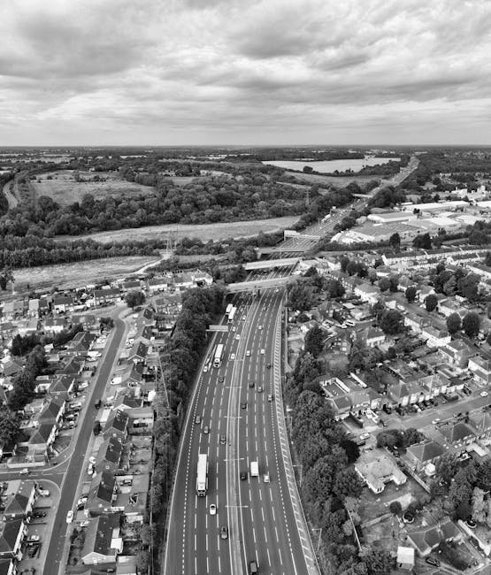 Aerial black and white photograph showing a residential neighbourhood with rows of houses and streets, adjacent to a busy multi-lane motorway with cars and trucks. To the right, there are trees and op