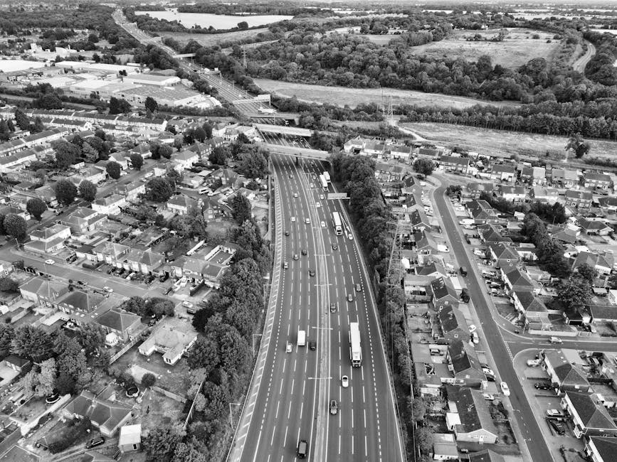 Aerial black and white photograph showing a residential neighbourhood with rows of houses and streets, adjacent to a busy multi-lane motorway with cars and trucks. To the right, there are trees and op