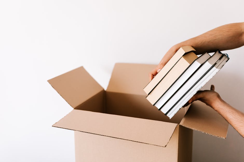 Inside a cardboard moving box, there is a black hardcover photo book featuring black-and-white images of various interior rooms, including a living room with a sofa, a dining area, and a fireplace, al