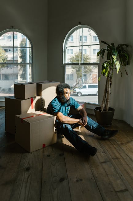 A man with curly hair and a beard, dressed in a blue shirt and dark trousers, is sitting on a wooden floor inside a room with large arched windows that allow natural daylight to illuminate the space. He is surrounded by several packed cardboard boxes, some sealed with red packing tape, indicating they are ready for moving or transport. Behind him, on the windowsill, there are additional cardboard boxes stacked, prepared for a home relocation or furniture transport process. The room features minimalist decor, including a tall potted plant near the window, adding a touch of greenery. Outside the windows, a street with parked cars and modern residential buildings is visible. The scene appears to be part of a professional removal or moving service setup, consistent with services offered by companies like Man with Van Thamesmead, supporting packing, loading, and logistics for house removals.
