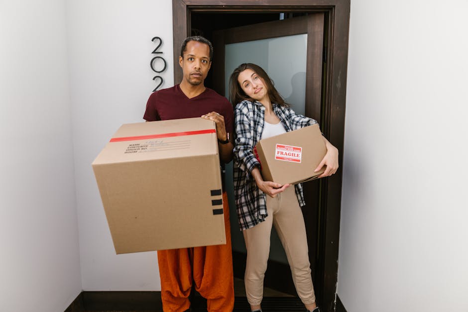 A man and a woman standing inside a residential doorway during a home relocation process, each holding cardboard boxes filled with household items. The man, dressed in an orange pair of trousers and a maroon t-shirt, holds a large box with black tape securing it, while the woman, wearing beige trousers and a plaid shirt, carries a smaller box labeled with 'FRAGILE' in red and white stickers. Behind them is a black door with a modern design, leading into a room with white walls. The scene captures the loading process involved in furniture transport and packing during a house removal, with the men with van Thamesmead providing professional moving services, including careful handling of boxes and loading belongings into a van nearby, visible through an open door or surrounding area, supporting efficient and secure relocation logistics.