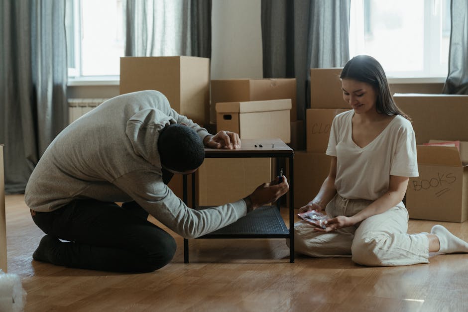 A man and a woman are inside a room during a home relocation, surrounded by cardboard boxes labeled with contents such as 'clothes' and 'books'. The man, dressed in a light grey sweater and dark trousers, is kneeling on the wooden floor and attaching a protective strap around a small black furniture trolley, which is positioned near a partly assembled black shelving unit. The woman, wearing a white T-shirt and light-coloured trousers, is sitting cross-legged on the floor holding a wrapped item, possibly packaging material or a small household object. In the background, several large cardboard boxes are stacked, some open or partially closed, with a large window providing natural light and grey curtains hanging on either side. The scene depicts the packing and furniture transport process typical of a flat-to-flat move, with attention to careful handling and protective preparations supported by a professional removals service like Man with Van Thamesmead.