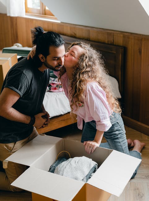 A woman and a young girl inside a room filled with cardboard moving boxes, some open and others sealed, during a home relocation process. The woman is kneeling on the wooden floor, organizing or packing items, while the girl, seated nearby, appears to be handling packing materials or toys. Part of a man is visible in the background, carrying a large cardboard box through a doorway, indicating the loading or unloading phase of furniture transport. The room is lit with natural light from a window, partially covered by curtains, and there are additional cardboard boxes stacked in the background. The scene captures the activity of packing and moving as part of a flat-to-flat removals service, with various packing supplies like cardboard boxes and bubble wrap present, highlighting the logistical steps involved in a professional house relocation. Man with Van Thamesmead specializes in efficient and organized furniture transport and packing during residential moves.