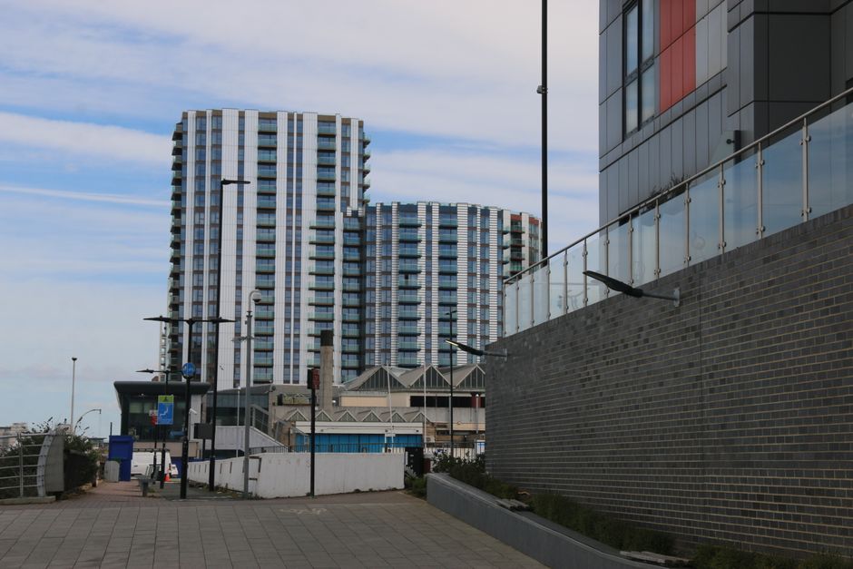 A daytime exterior view of a modern urban area showing high-rise residential buildings with glass balconies in the background, adjacent to a commercial or office building with a brick and glass facade