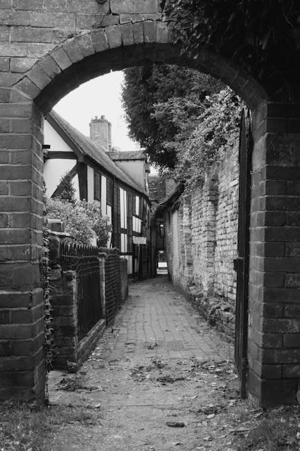 A narrow cobbled alleyway viewed through a brick archway, leading between residential buildings and a stone wall on the right. The alley is used as a loading area for a home removal process with visible packing materials, including cardboard boxes, plastic wrapping, and moving blankets, placed near a parked van. The residential buildings on the left have traditional timber-framed facades with windows, while the stone wall on the right appears weathered. A man with a van is engaged in furniture transport, possibly unloading or loading items as part of a house relocation along the Thamesmead West route. The scene is set during daylight with natural lighting highlighting the textures of the brick, stone, and cobbles, capturing a typical urban environment for professional removals or packing and moving services by Man with Van Thamesmead.