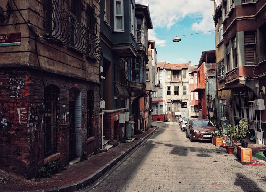 A narrow cobbled alleyway viewed through a brick archway, leading between residential buildings and a stone wall on the right. The alley is used as a loading area for a home removal process with visible packing materials, including cardboard boxes, plastic wrapping, and moving blankets, placed near a parked van. The residential buildings on the left have traditional timber-framed facades with windows, while the stone wall on the right appears weathered. A man with a van is engaged in furniture transport, possibly unloading or loading items as part of a house relocation along the Thamesmead West route. The scene is set during daylight with natural lighting highlighting the textures of the brick, stone, and cobbles, capturing a typical urban environment for professional removals or packing and moving services by Man with Van Thamesmead.
